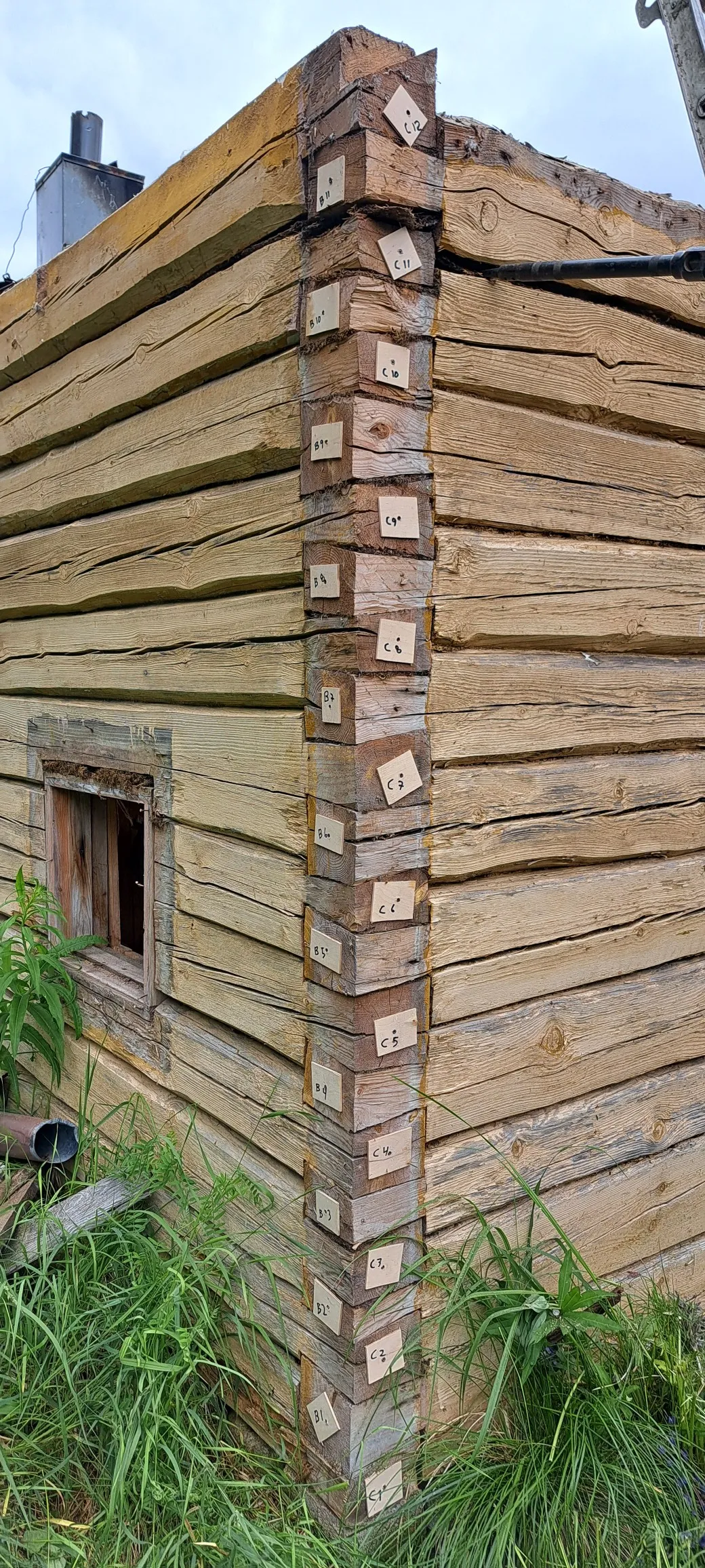 Lähikuva vanhasta hirsitalosta, jonka nurkassa on paperilappuja merkinnöillä. Hirsien väri on kellertävä ja valaistus on luonnollinen.