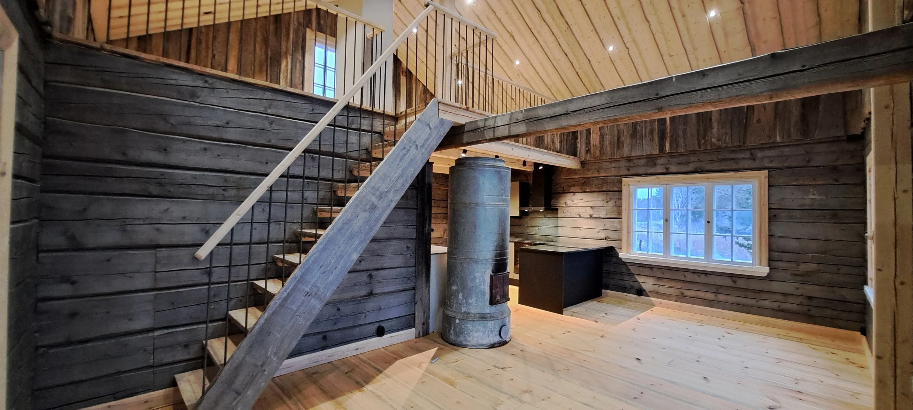 Sisätila hirsitalossa, jossa on puuportaat, metallikaide, puulattia, puuseinät, ikkuna ja vanha metallinen kamiina.