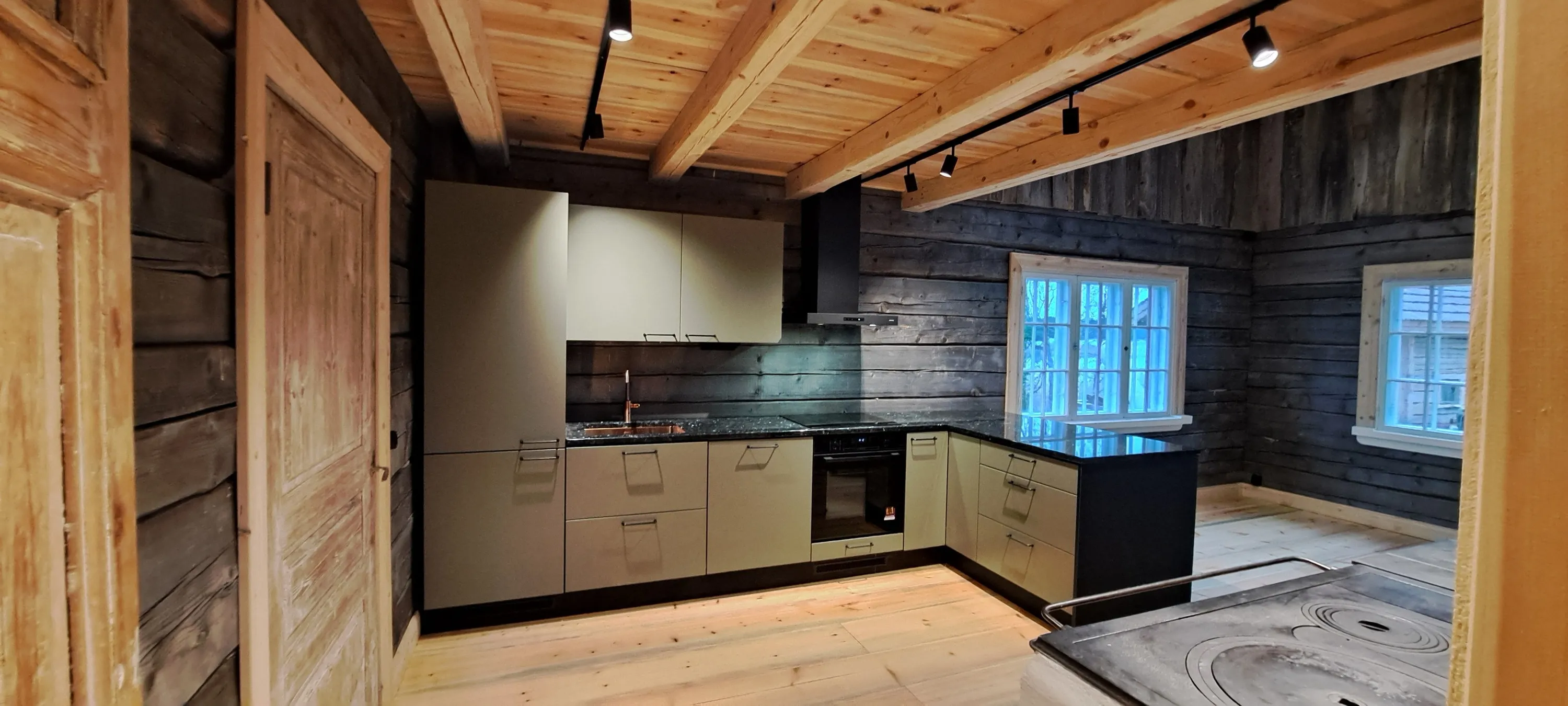 Moderni keittiö hirsitalossa, jossa on vaaleat kaapit, musta työtaso ja puinen katto, valaistu kohdevaloilla.