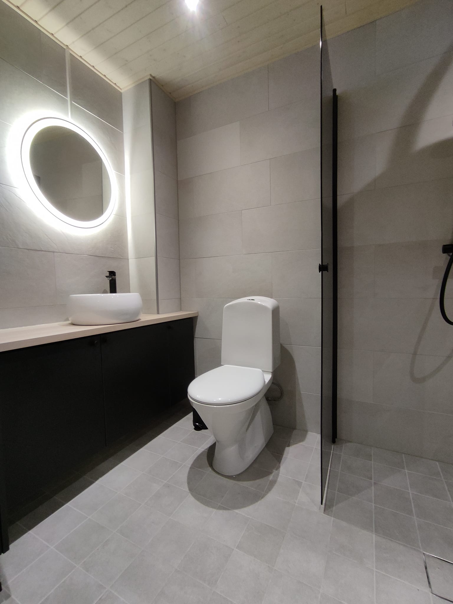 Moderni kylpyhuone, jossa on pyöreä peili, valkoinen pesuallas, musta allaskaappi, valkoinen wc-istuin ja harmaat laatat.