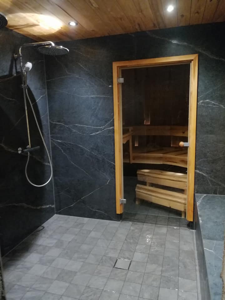 Kylpyhuoneen sisustus, jossa on suihku, laattalattia, tummat marmoriseinät ja puinen sauna lasiovella.