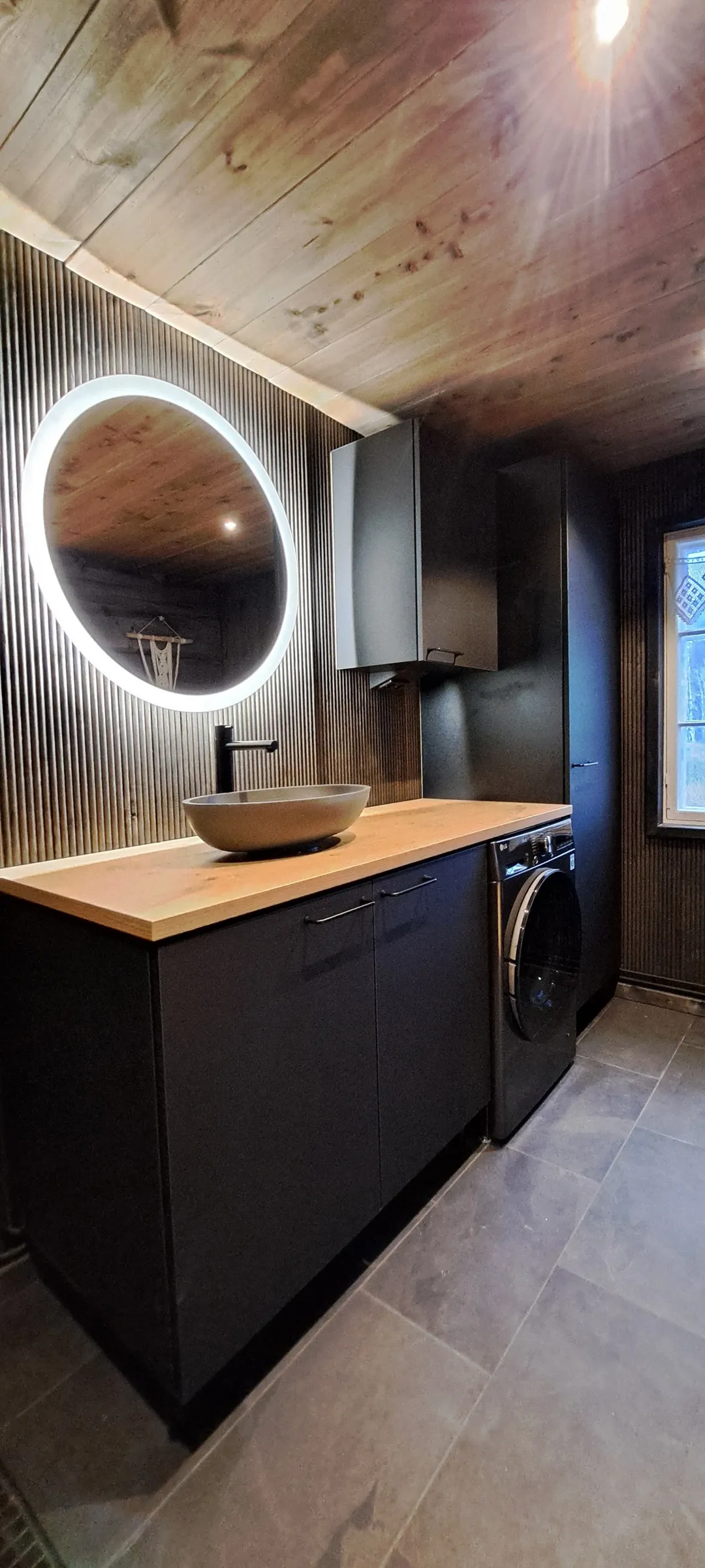 Moderni kylpyhuone, jossa on puukatto, pyöreä peili, mustat kaapit, puinen työtaso ja harmaa pesuallas.