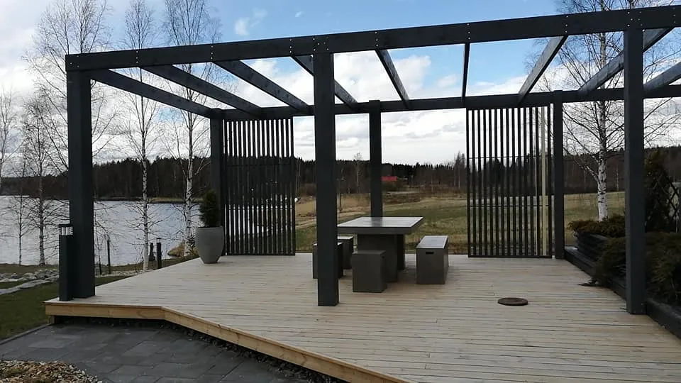 Ulkona oleva puinen terassi, jossa on harmaa pergola, pöytä ja penkit, sekä näkymä järvelle ja metsään.