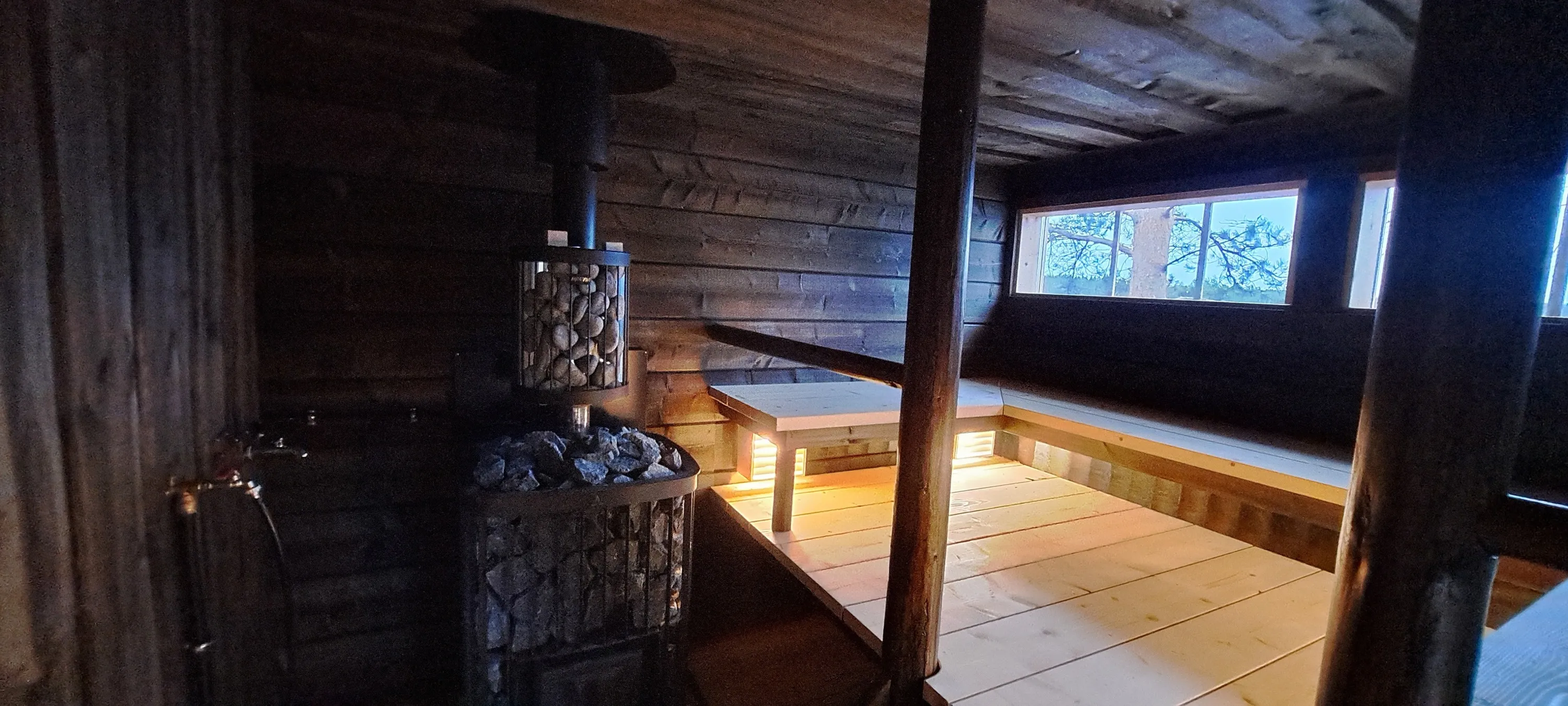 Tumma puusauna, jossa kivikasa, puupaneelit, ikkunat ja valaistus luovat lämpimän tunnelman.