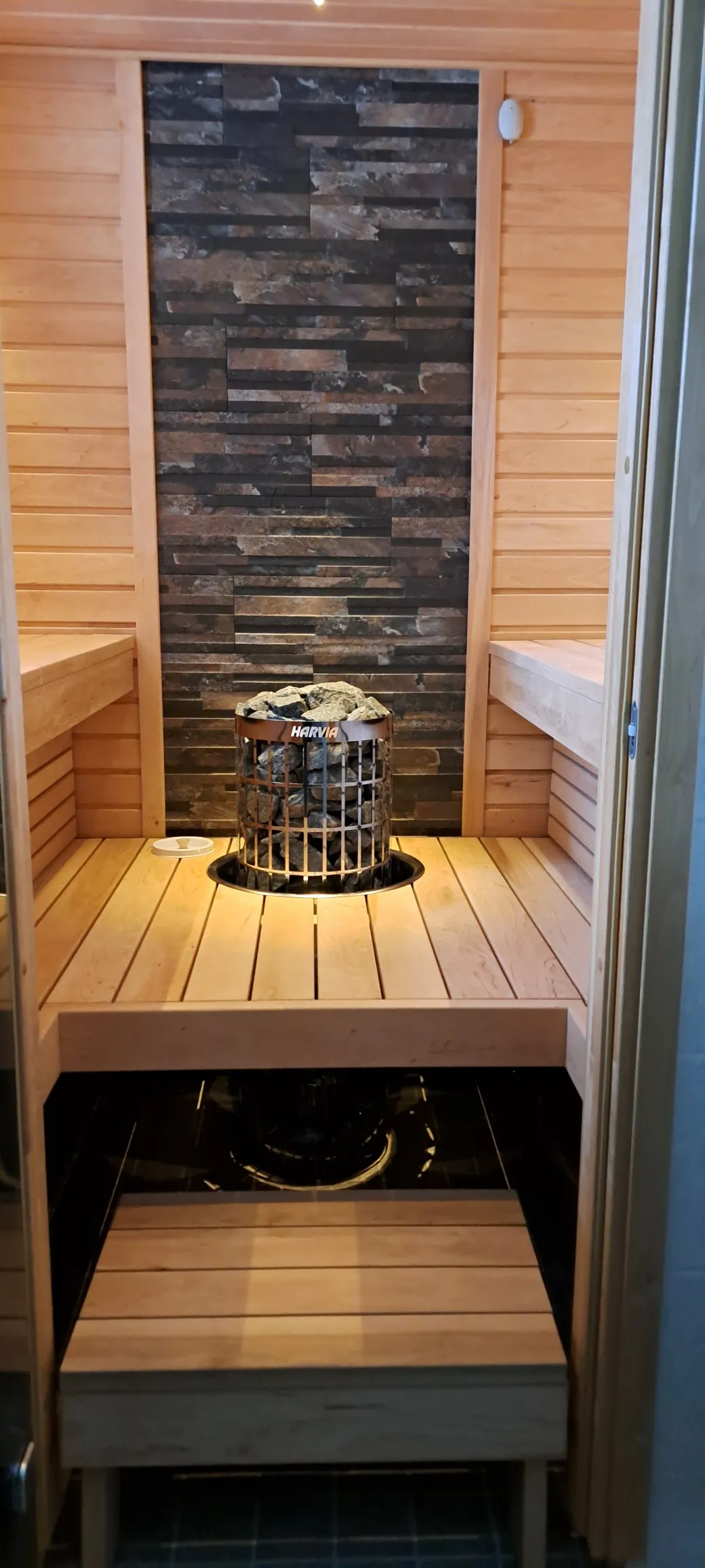 Sauna, jossa on puupanelointi, kiviseinä ja Harvia-kiuas täynnä kiviä, valaistus luo lämpimän tunnelman.