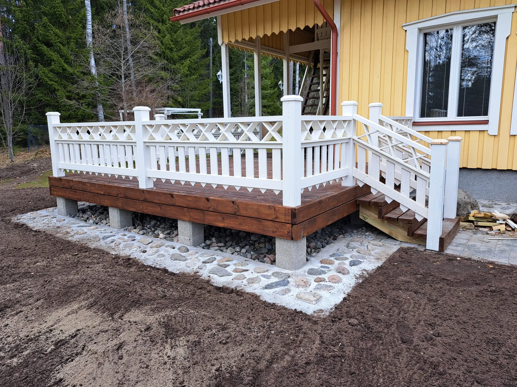 Ulkoterassi valkoisella kaiteella, ruskealla puukannella ja kivijalalla, aurinkoisessa säässä.